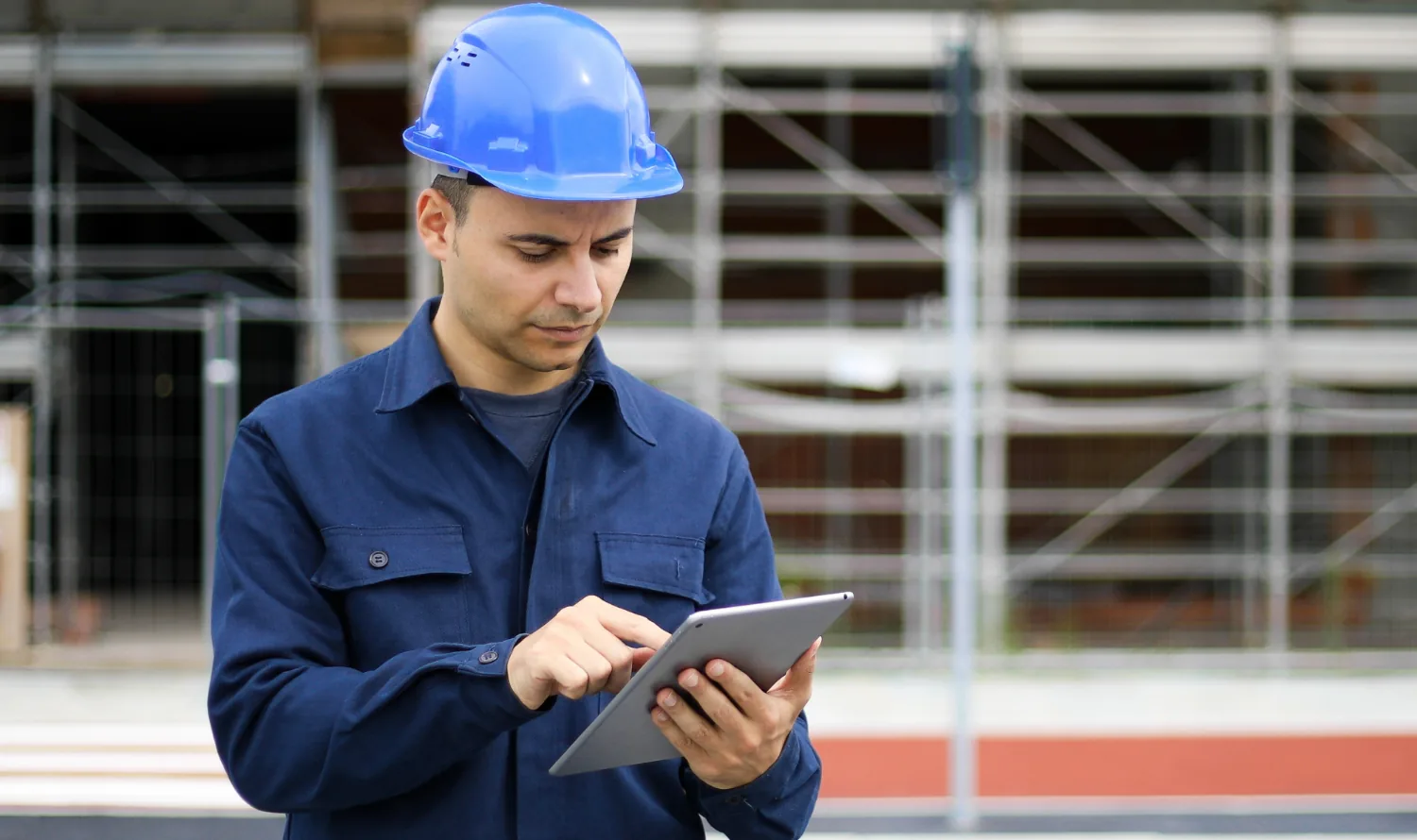 Técnico con casco azul revisando una tableta frente a un andamio